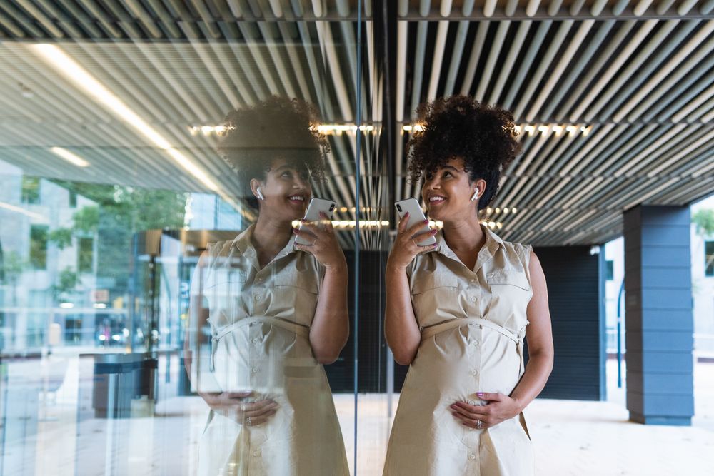 Smiling Black pregnant woman using smartphone and wireless earphones