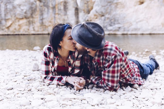 Positive woman and man resting and kissing on stone shore near river