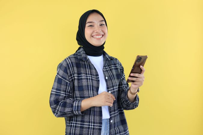 Portrait of Smiling Young Asian Woman in Blue Checkered Shirt and Hijab using Mobile Phone