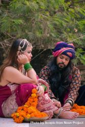 Indian man with long hair wearing turban sitting beside woman in colorful dress on floor outdoor 4BYRX5