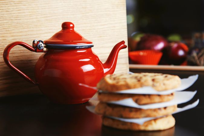Shiny red tea pot on wooden counter with pile of cookies