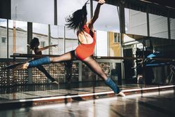Back view of young slim ballerina jumping above ground in studio flexing legs.