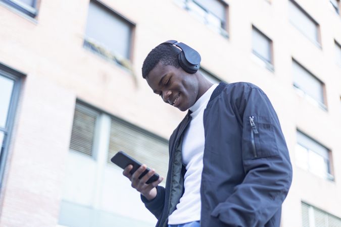 Smiling Black man leaning outside, wearing headphones and looking down at his phone