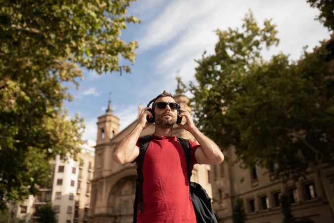 A young, traveling man, wearing sunglasses, puts on headphones to listen to music during a walk through a city in Spain