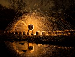 Person with burning circle on stones between water near hills at night