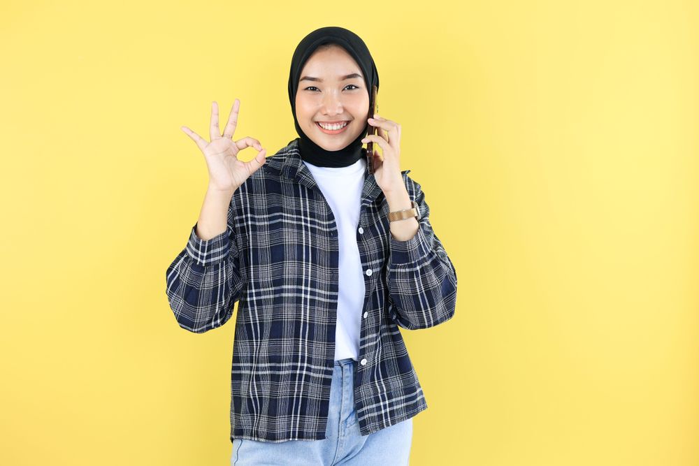 Young Asian Woman Wearing Hijab Talking on Cell Phone with Friend, Looking at Camera While Posing Okay.