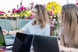 Two smiling friends comparing their notes and chatting about what was written on the laptop.