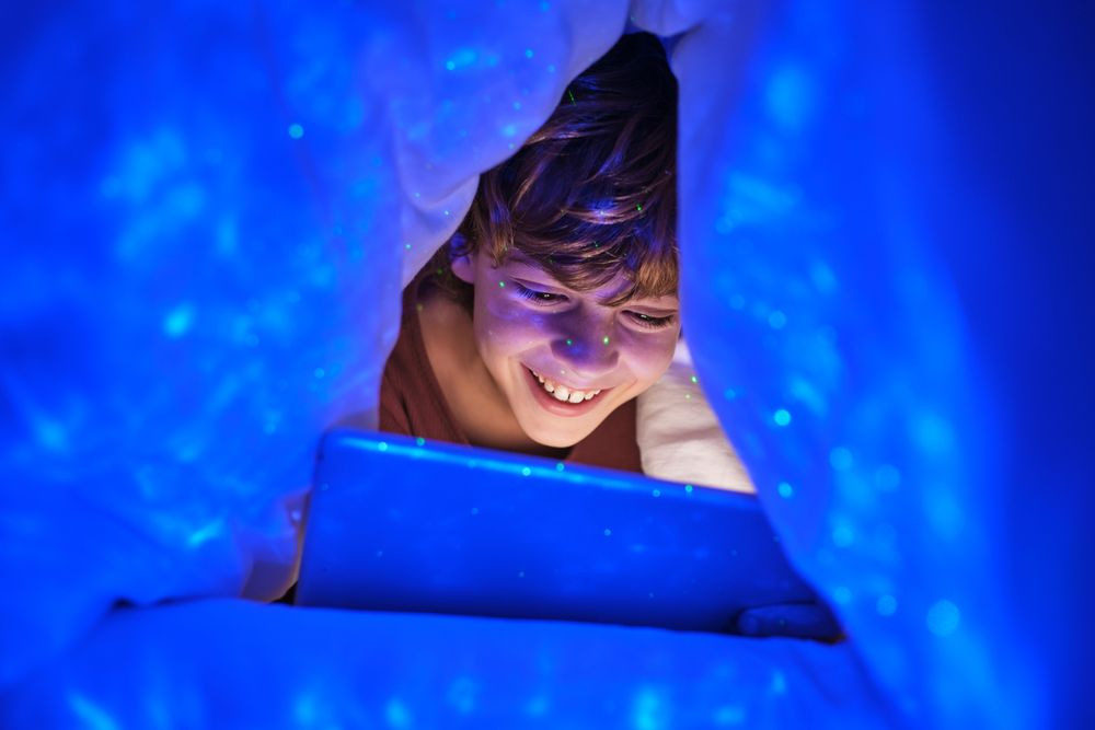 Cheerful boy browsing tablet under blanket