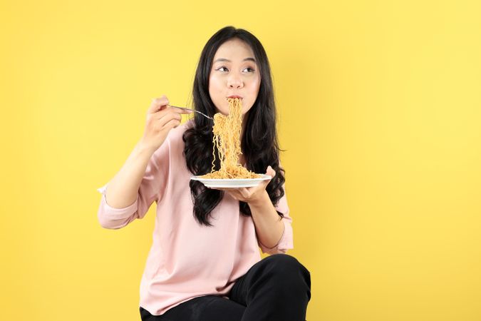 Happy Young Asian Woman Sitting on a Chair Wearing Pink Shirt, Eating Noodles on White Plate using Fork.