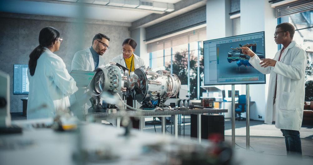 Diverse Professional Jet Engine Researchers Discussing Work, Using Laptop Computer and Checking Blueprints in Motor Manufacturing Facility.