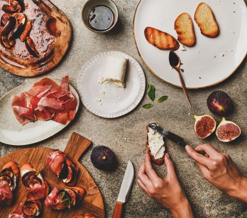 Crostinis with prosciutto, goat cheese and grilled figs, with hands spreading cheese, square crop