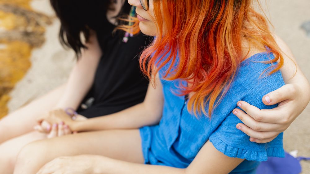 A young female hugs and comforts her partner as she hugs her shoulder and shakes her hand