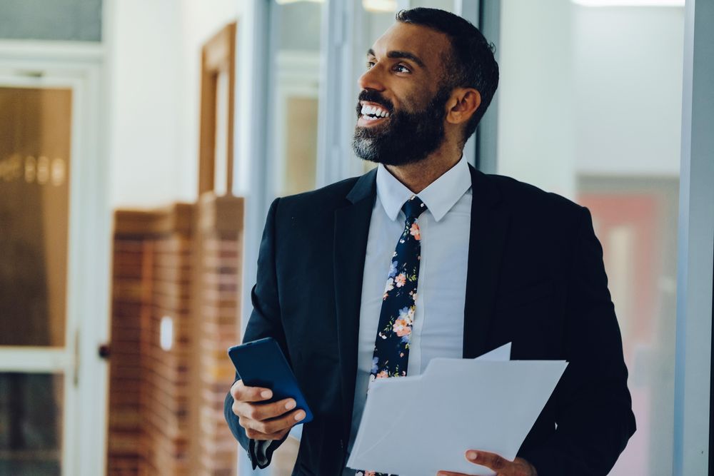 Happy businessman in suit and tie with papers standing in office while holding smart phone