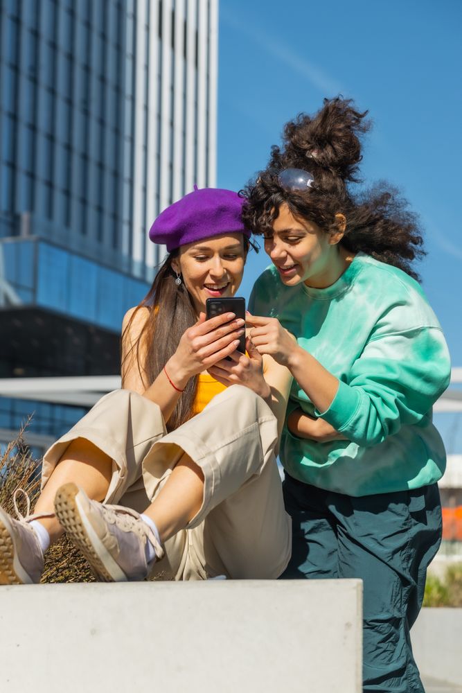 Two Young Women Sharing A Laugh While Looking At A Smartphone In A City Setting, Showcasing Friendship And Technology.