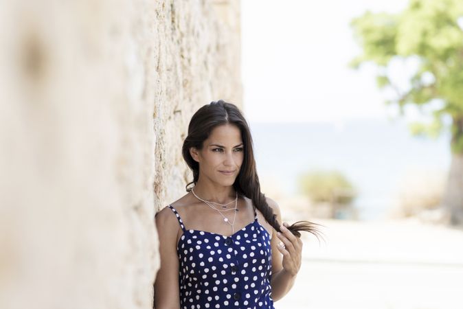Latina woman in blue dotted dress leaning on wall outside and looking away playing with hair