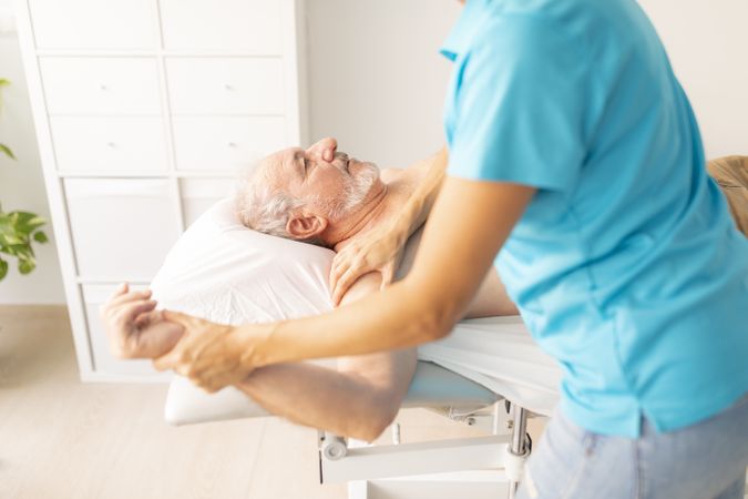 An older man, lying on the table in a physiotherapy clinic, during a therapy session with his physio for shoulder mobility
