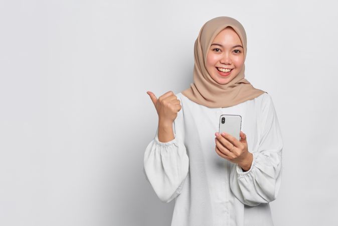 Asian Muslim woman in a bright studio shoot holding cell phone and gesturing to side with thumb