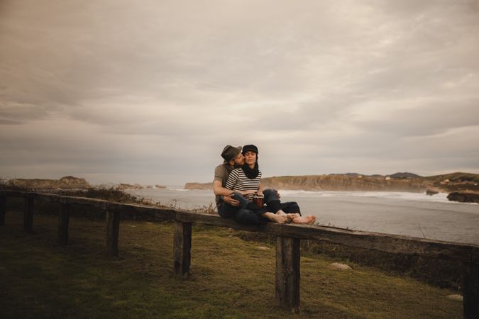 Young guy in hat near elegant woman playing on hand drum in cap on seat near coast of sea and cloudy sky