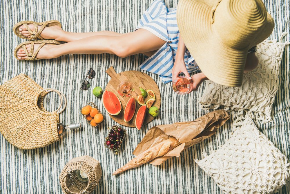 Woman on picnic blanket with  thatched bag with glass and bottle of rose, baguette, watermelon