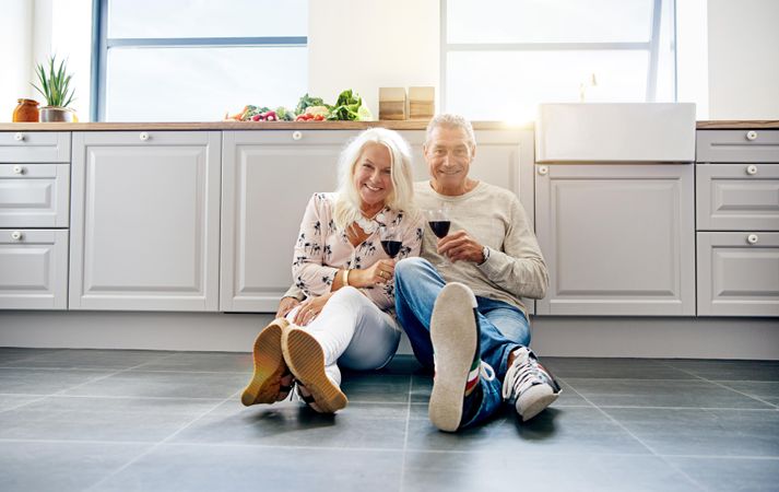 Happy older couple looking at camera and smiling while enjoying wine on kitchen floor