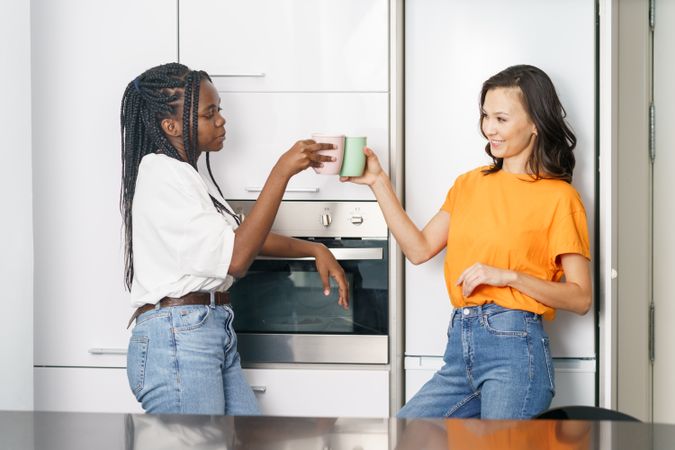 Two women talking while leaning on the fridge while raising their mugs