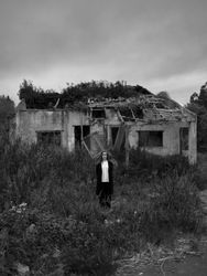 A young woman stands alone in a rugged setting, surrounded by tall grasses and an abandoned, crumbling building in the background