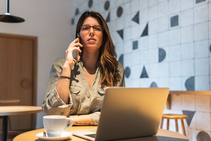Woman speaking on smartphone against laptop in cafeteria