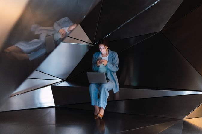 Woman with laptop browsing smartphone in futuristic studio