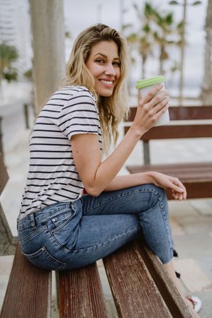 Happy woman with disposable cup of coffee sitting on street bench