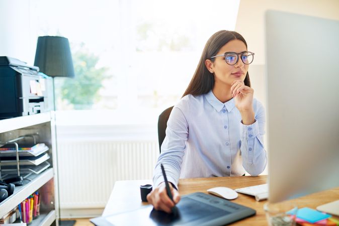 Female graphic designer using stylus on digital drawing pad and looking at monitor in her office