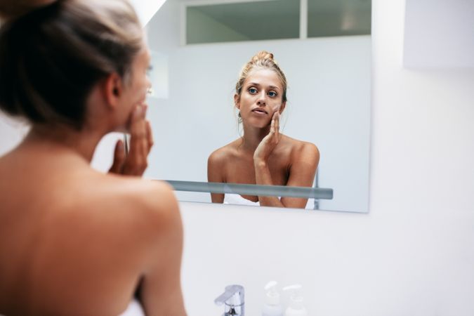 Beautiful young woman in bathroom looking in to mirror and applying moisturizer on face