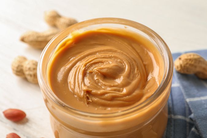 Glass jar with peanut butter, peanut and kitchen towel on wooden background, closeup and space for text