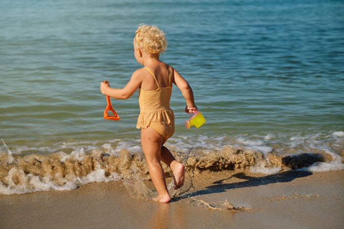 Blonde child in yellow swimsuit going to sea with toys