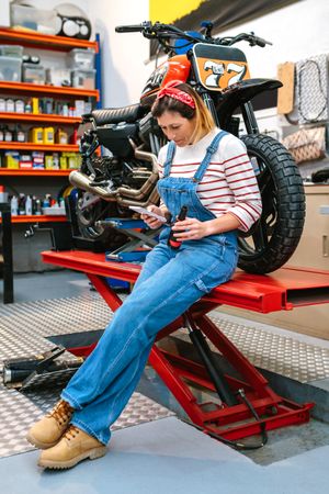 Mechanic woman looking phone and holding beer sitting over platform with motorcycle on factory