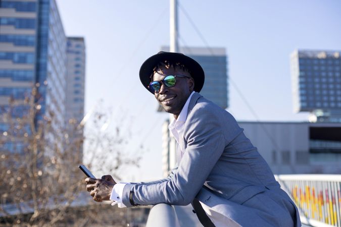 Smiling Black man wearing hat and sunglasses leaning on a metallic fence while using a mobile phone outdoors in a sunny day
