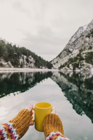 Person with cup near wonderful lake between hills in snow and cloudy sky