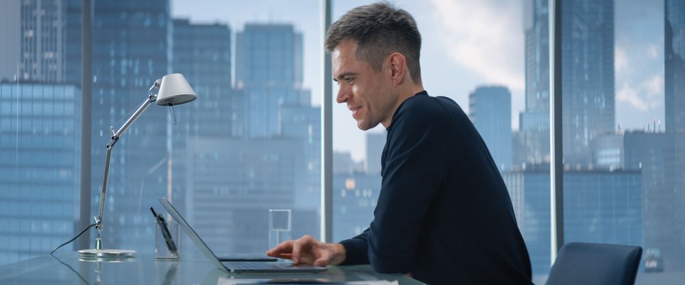 Confident Businessman Sitting at a Desk in Modern Office, Using Laptop Computer, Next to Window with Big City with Skyscrapers View.