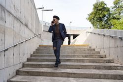 Young Latino man wearing a fashionable hat and sunglasses, walking down the stairway while texting on his smartphone