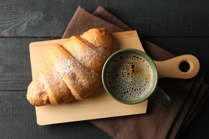 Board with croissant and mug of coffee on dark wooden background, top view