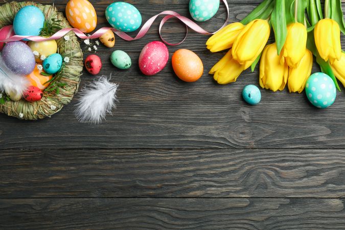 Nest with olorful easter eggs, flowers and feathers on wooden background. Festive tradition. Space for text. Top view