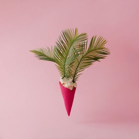 Palm leaves emerging from vanilla ice cream in pink waffle cone on pink pastel background