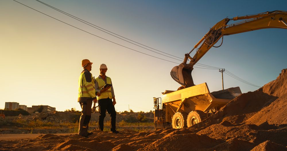 Real Estate Investor With Tablet And Project Manager Talking On Construction Site Of Apartment Building. Colleagues Wearing Hard Hats And Watching Heavy Machinery Working. Sunny Day