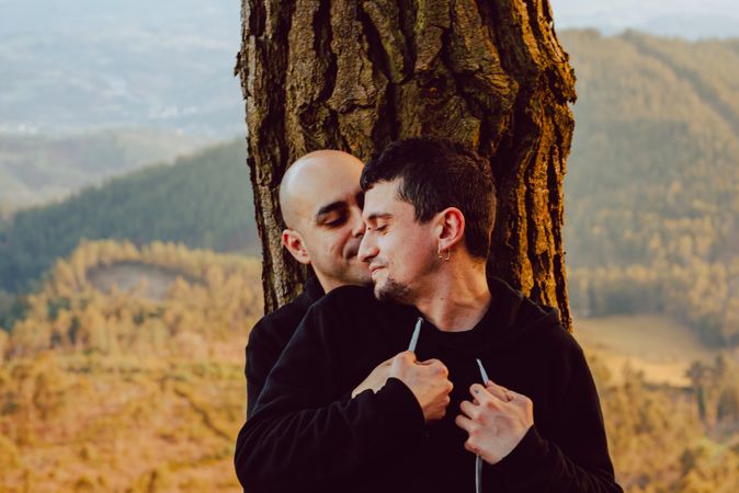 Smiling men hugging near tree in forest on hill
