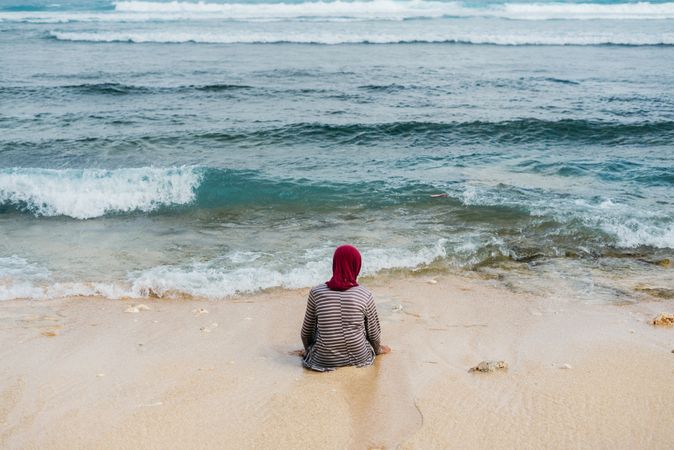 Back of Muslim woman sitting at water's edge on Bali beach