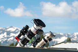 Telescopes on observatory rooftop with mountains and blue sky.