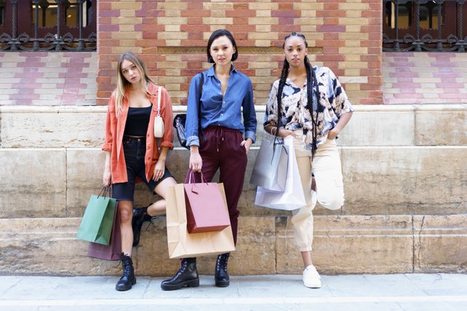 Three proud women posing with shopping bags in front of brick wall
