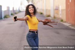 Smiling woman spinning around in center of street holding bag and phone 0LROP4