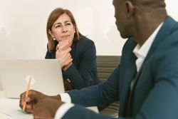 In a bright office, a mature woman with long, straight brown hair is listening in deep concentration