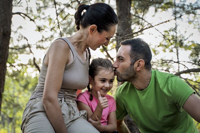 Parents kissing smiling daughter in forest during family bonding time outdoors