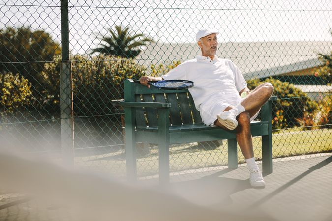 Older man sitting on a bench beside a tennis court and relaxing